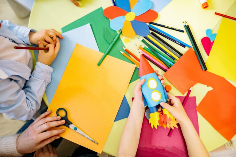 Imagen de manos de niños y su instructora con folios y lápices de colores